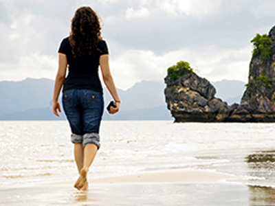 A woman walking away from the camera along a beach. Rocky sea stacks and mountains are in the distance