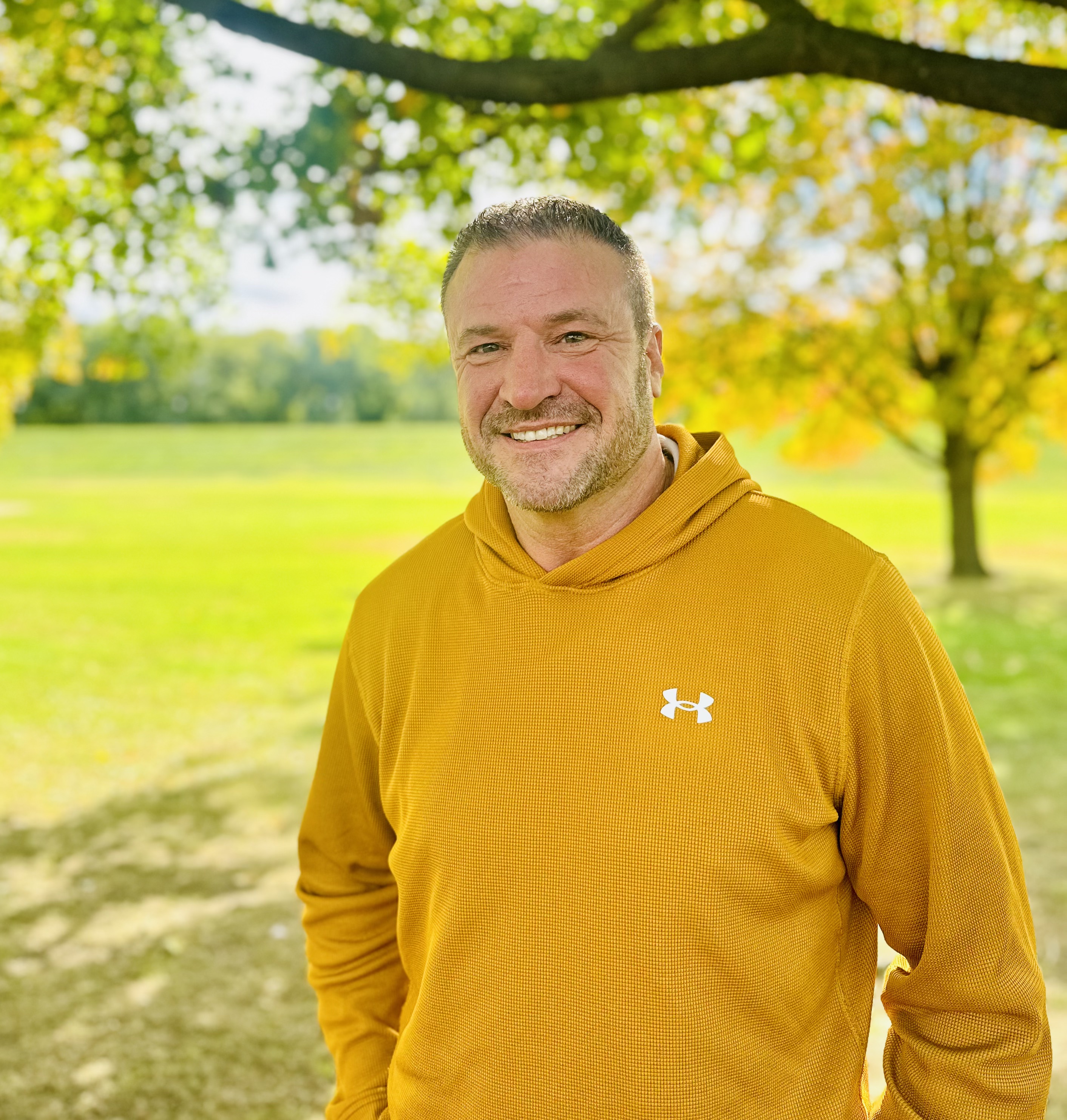 Chris Neyman standing in a park and smiling at the camera wearing a yellow sweatshirt