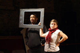 A woman and man on a dark stage. The woman is holding up a cardboard television frame, and the man is standing with his head in the frame and speaking.